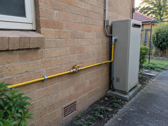 Yellow gas piping on a brick exterior wall beside a grey appliance and garden path in Normanhurst